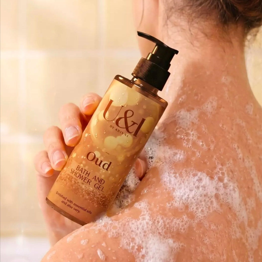 U&I Oud bath and shower gel in a 200ml amber bottle held against a shoulder with soap lather in a warm bathroom setting.