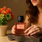U&I In My Peach Girl Era perfume bottle with peach-toned liquid, held by a woman near a small potted flower plant.